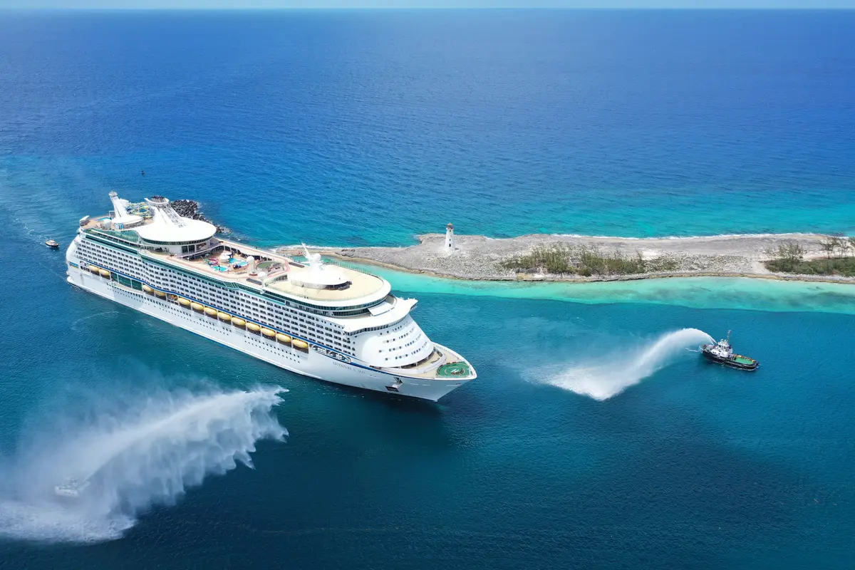 Luftaufnahme der Adventure of the Seas im tropischen Meer. Ein Schlepper spritzt Wasser, eine Insel mit Leuchtturm ist sichtbar.