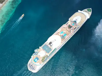 Luftaufnahme der Adventure of the Seas in türkisblauem Wasser nahe einer Küste. Man sieht Pools, Sportdeck und ein kleines Begleitboot.
