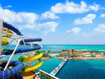 Blick von der Adventure of the Seas auf eigene bunte Wasserrutschen. Eine tropische Insel mit Wasserpark, Stränden und bunter Pier liegt im türkisblauen Meer.