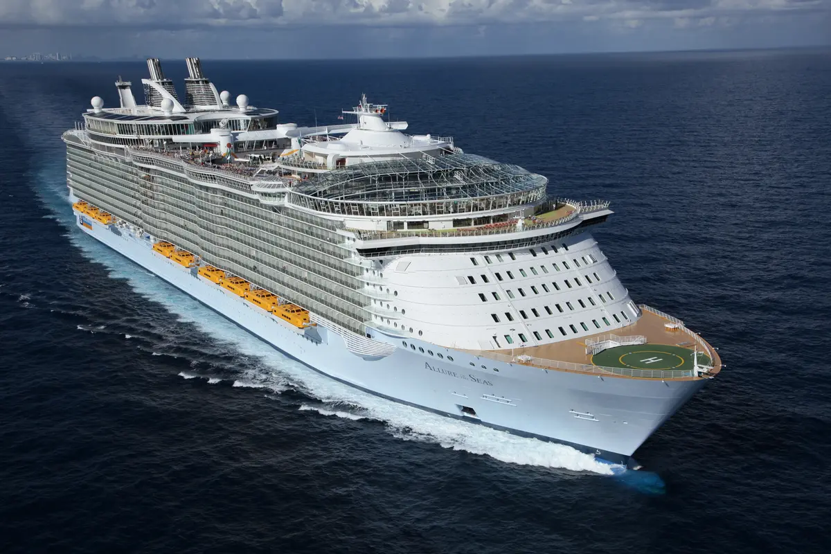 Luftaufnahme der Allure of the Seas auf blauem Meer. Viele Balkone, orange Rettungsboote, Glaskuppel, Helipad und eine Skyline am Horizont.