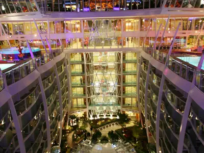 Der Blick von oben zeigt das imposante, mehrstöckige Atrium der Allure of the Seas. Man sieht zahlreiche Kabinenbalkone, die das offene Zentrum umrahmen. Unten befindet sich eine begrünte Parklandschaft mit Wegen und Pflanzen. Höhere Decks beherbergen Geschäfte und Pools.