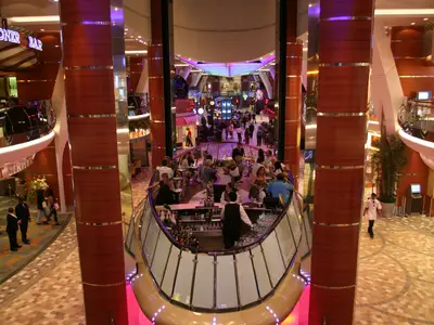 Blick in die belebte Promenade der Allure of the Seas. Eine Bar mit Gästen und Barkeeper ist im Vordergrund. Dahinter sind Geschäfte und viele Passagiere auf mehreren Ebenen zu sehen.