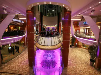 Innenansicht der Allure of the Seas: Das Atrium mit zentraler Bar und Gästen, einem lila beleuchteten Brunnen und der Schooner Bar auf einer oberen Ebene.