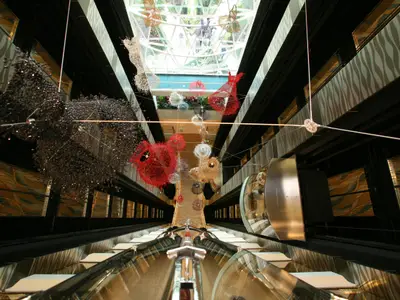 Vertikale Aufnahme des mehrstöckigen Atriums an Bord der Allure of the Seas, geschmückt mit farbigen, schwebenden Kunstobjekten und gläsernen Panoramaaufzügen. Eine Glasdecke ist oben sichtbar.
