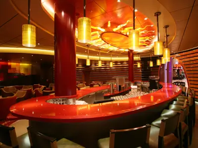 Eine elegante Bar an Bord der Allure of the Seas. Rote Theke, goldene Hängelampen, kunstvolle orange Decke mit flammenden Mustern. Bequeme Sessel im Hintergrund.