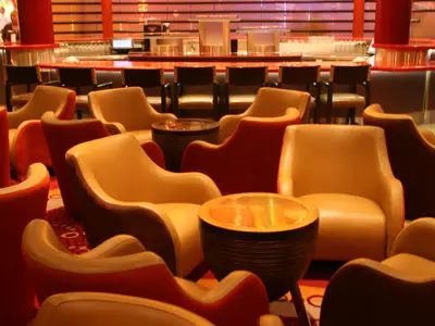 Sicht in eine moderne Lounge auf der Allure of the Seas mit beigefarbenen und roten Sesseln an runden Tischen. Im Hintergrund eine beleuchtete Bar mit Hockern.