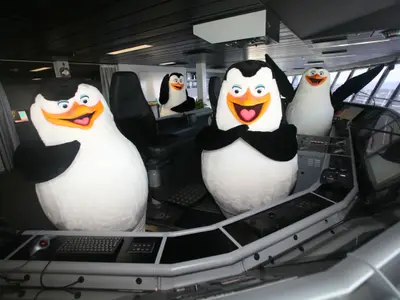 Vier Pinguin-Maskottchen auf der Kommandobrücke der Allure of the Seas. Sie sind zwischen den Bedienkonsolen und Stühlen positioniert, mit Blick nach draußen durch die Fenster der Brücke.