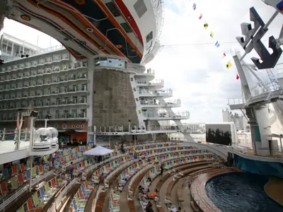 Blick auf das Heck der Allure of the Seas: Das offene AquaTheater mit Pool und gestuften Sitzreihen ist zentral. Darüber erheben sich zahlreiche Balkonkabinen und eine Kletterwand. Am Himmel wehen maritime Signalflaggen.