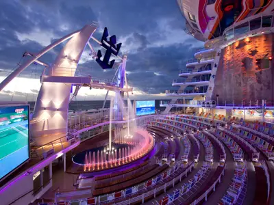 Das abendliche AquaTheater der Allure of the Seas. Man sieht farbige Fontänen, leere Sitzreihen, große Bildschirme und eine Felsenkletterwand.