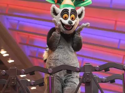Ein lebensgroßer King Julian-Charakter aus Madagascar mit grauem Fell, orangefarbenen Augen und einem breiten Lächeln. Er steht auf einer dunklen Plattform mit erhobenen Händen. Im Hintergrund sind diffuse rote und violette Lichter zu sehen.