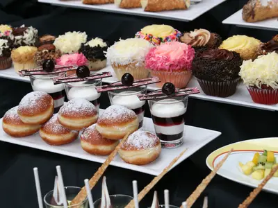 Ein Tisch voller Desserts, darunter eine Platte mit Puderzucker-Donuts, mehrere Gläser mit Schichtdesserts und im Hintergrund eine Reihe bunter Cupcakes auf einem weißen Tablett.