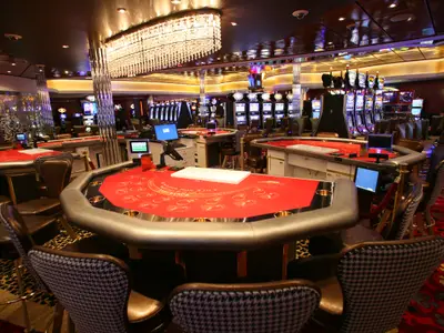 Blick in das elegant beleuchtete, unbesetzte Casino der Allure of the Seas. Mehrere rote Spieltische und Reihen von Slotmaschinen sind sichtbar, überragt von einem großen Kronleuchter.