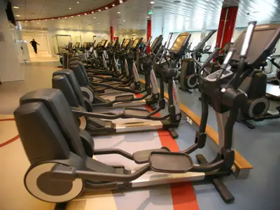 Eine Innenansicht des weitläufigen Fitnessstudios an Bord der Allure of the Seas, das zahlreiche moderne Crosstrainer in Reihen zeigt. Eine Person läuft im hellen Hintergrund.