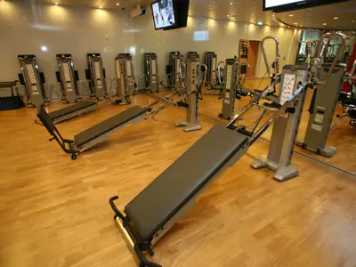 Fitnessraum auf der Allure of the Seas: Mehrere Total Gym Geräte auf hellem Holzboden. Eine Spiegelwand reflektiert Trainingsmaschinen und einen TV-Bildschirm.