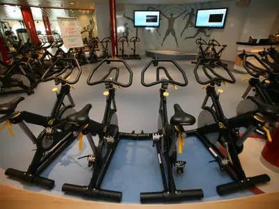 Das Fitnessstudio der Allure of the Seas, ausgestattet mit Spinning-Rädern und weiteren Geräten. Bildschirme und Wandmotive zeigen Athleten.