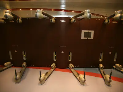 Modernes Technogym Kinesis-Krafttrainingsgerät an einer braunen Holzwand im Fitnessbereich. Mehrere bewegliche Arme mit Griffen und ein kleiner Bildschirm sind zu sehen. Der Boden ist hellgrau mit einem orangefarbenen Streifen.