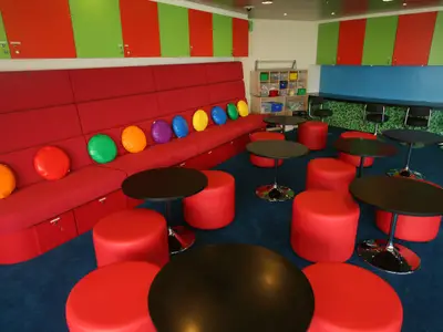 Ein lebhaft gestalteter Kinderbereich mit roten Sitzbänken, die mit bunten Kissen in verschiedenen Farben dekoriert sind. Mehrere rote Hocker und schwarze Tische stehen auf einem blauen Teppich. Im Hintergrund sind grüne und rote Schränke sowie ein Regal mit Spielboxen zu sehen. Ein spielerisches Interieur.