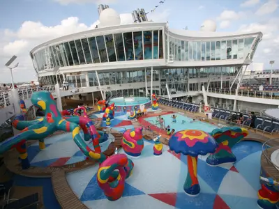 Blick auf den bunten H2O Zone Wasserpark an Deck der Allure of the Seas, mit farbenfrohen Spielfiguren, einem Pool voller Badegäste, Whirlpools und Liegestühlen unter klarem Himmel.