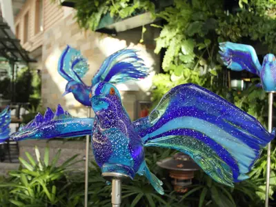 Nahaufnahme mehrerer leuchtend blauer Glasvogel-Skulpturen mit weißen Sprenkeln und orangen Augen. Sie stehen auf Stangen vor einem Hintergrund aus grünen Pflanzen und einem Gebäude.