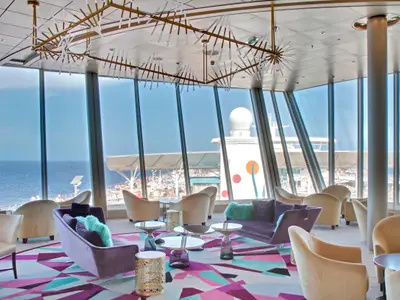 Allure of the Seas: Moderne Lounge, bunter Teppich, Möbel. Große Fenster mit Blick auf Meer, Schiffsaufbauten, Radom, bemalter Schornstein.