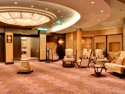 Lounge auf der Allure of the Seas: Großer Kronleuchter über hellen Sesseln, Sofa und Tischen auf gemustertem Teppich. Holzvertäfelung und warme Beleuchtung schaffen eine elegante Umgebung.