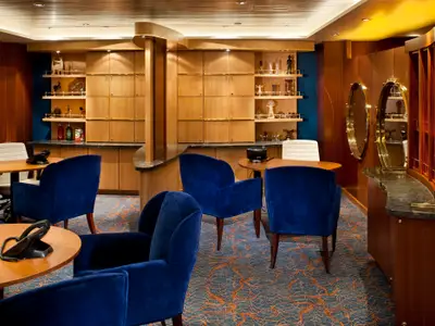 Auf der Allure of the Seas: ein Loungebereich mit blauen Sesseln, Holztischen und Telefonen. Die Holzwände zeigen Regale, Dekorationen und ovale Spiegel. Blauer Teppich mit Wellenmuster.