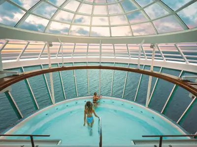 Zwei Frauen in einem Whirlpool an Bord der Allure of the Seas, umgeben von großen Glasfenstern, die einen weiten Blick auf das offene Meer und den Himmel bieten.