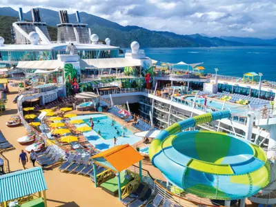 Das Oberdeck der Allure of the Seas zeigt mehrere Pools, eine Wasserrutsche, zahlreiche Liegestühle und Passagiere. Berge und Meer im Hintergrund unter leicht bewölktem Himmel.