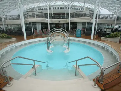 Das Solarium auf der Allure of the Seas zeigt einen großen, runden Pool mit Wasserspiel und Treppen. Umgeben ist er von Holzdecks und mehreren Lounge-Ebenen unter einem transparenten Dach.