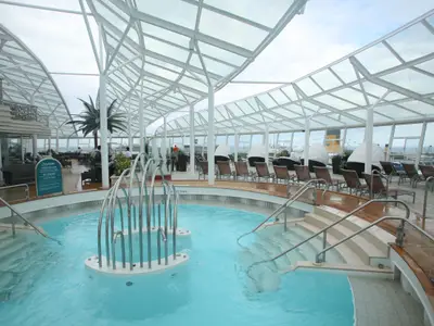 Poolbereich im Solarium der Allure of the Seas. Ein großer Pool mit Wasserfontäne unter einem Glasdach, umgeben von Liegestühlen. Durch die Fenster ist das Meer sichtbar.
