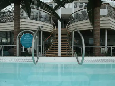 Ein Adults Only Whirlpool mit Metallgriffen ist auf der Allure of the Seas im Vordergrund zu sehen. Dahinter führen geschwungene Treppen, von Palmen eingerahmt, zu oberen Decks mit Glasbalustraden und Lounge-Bereichen.