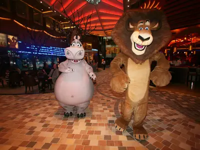 Zwei Maskottchen, Gloria das Nilpferd und Alex der Löwe aus Madagascar, stehen auf der Royal Promenade der Allure of the Seas. Im Hintergrund sind Passagiere und eine Bar zu sehen.