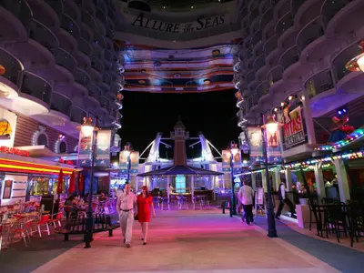 Nächtliche Szene auf der Royal Promenade der Allure of the Seas. Beleuchtete Restaurants und Geschäfte säumen den Weg, Menschen flanieren. Im Vordergrund ein Paar.