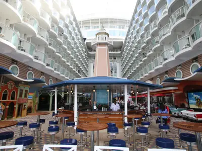 Außendeck der Allure of the Seas mit zentraler Bar und Leuchtturm, flankiert von Balkonkabinen. Bunte Gebäude und ein großer Bildschirm sind sichtbar.