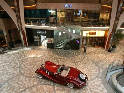 Eine Ansicht der Royal Promenade auf der Allure of the Seas zeigt einen roten Oldtimer. Geschäfte wie Guess und eine Wendeltreppe sind sichtbar, oben die Focus Photo Gallery.