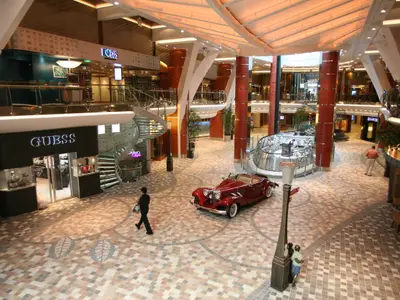 Blick von oben auf die Royal Promenade der Allure of the Seas: GUESS-Geschäft, Wendeltreppe, roter Oldtimer und Geschäfte auf mehreren Ebenen.