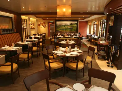 Innenansicht eines Restaurants auf der Allure of the Seas. Gedeckte Tische, Holzsessel, gelbe Polster, warme Beleuchtung und eine Landschaftsmalerei.