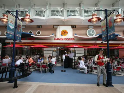Blick auf den Boardwalk des Schiffs Allure of the Seas mit dem Johnny Rockets Restaurant, Gästen im Außenbereich und einer Figur eines Akkordeonspielers. Oben sind Balkonkabinen sichtbar.