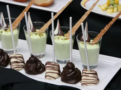 Nahaufnahme von fünf Gläsern mit grünlichem Dessertpüree und schokoladenen oder knusprigen Stäbchen. Davor liegen fünf Schoko-Pops, teils dunkel, teils hell glasiert, auf einem weißen Tablett.