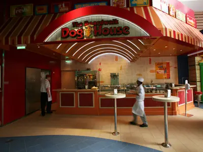 Blick in das Boardwalk Dog House auf der Allure of the Seas, einer Hotdog-Bar an Bord. Zwei Mitarbeiter, einer am Eingang, einer an der Theke, sind im farbenfrohen Innenraum zu sehen.
