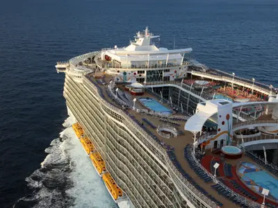 Luftaufnahme der Allure of the Seas auf dem Meer. Man sieht die Oberdecks mit mehreren Pools, Whirlpools, Sonnendecks mit Liegen, sowie Reihen von gelben Rettungsbooten an der Bordseite.
