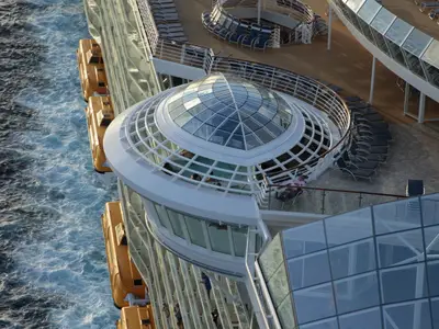 Blick von oben auf die Allure of the Seas. Man sieht die Schiffsseite mit Rettungsbooten im Meer, dazu eine Glaskuppel und Decks mit Liegestühlen und Personen.