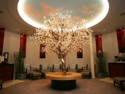 Ein geräumiger, elegant gestalteter Spa-Loungebereich mit einem großen, blühenden Kirschblütenbaum als Mittelpunkt. Bequeme Sessel und eine Deckenmalerei mit Wolken ergänzen das entspannte Ambiente. Eine Getränkestation ist seitlich sichtbar.