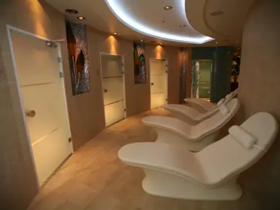 Ein heller Spa-Ruhebereich mit vier weißen, geschwungenen Liegen auf dem Boden. Entlang einer leicht gekrümmten Wand befinden sich mehrere Milchglastüren, die zu verschiedenen Kabinen führen. Die Wände sind mit beigen Fliesen und dekorativen Mosaikstreifen versehen. Indirekte Beleuchtung von der Decke.