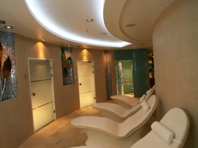 Moderner Entspannungsbereich an Bord der Allure of the Seas mit weißen Lounge-Liegen, hellen Wänden, Mosaikakzenten, Glastüren und einem grün gefliesten Duschbereich.