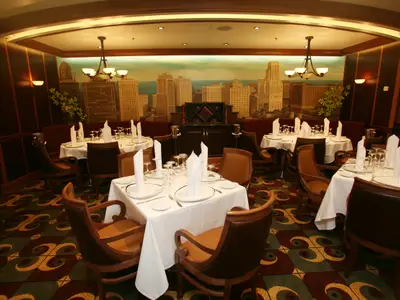 Innenansicht eines eleganten Restaurants an Bord der Allure of the Seas. Mehrere Tische sind mit weißen Tischtüchern, Tellern und Gläsern gedeckt. Braune Sessel und ein Wandgemälde mit Stadtkulisse prägen den Raum.