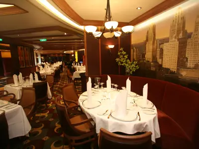 Ein Restaurant auf der Allure of the Seas mit vielen gedeckten Tischen und eleganter Einrichtung. Eine rote Sitzbank vor einem Stadt-Wandbild. Kronleuchter und Holzdetails prägen den Raum.