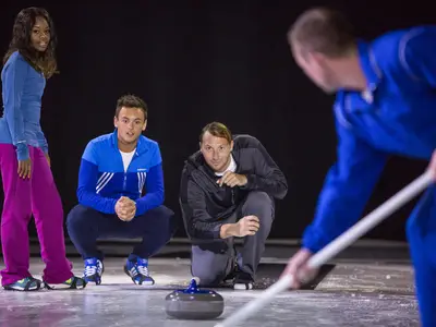Athleten Tom Daley und Ian Thorpe beobachten ein Curling-Spiel auf Eis. Eine Person fegt, andere sehen zu. Pressefoto für die Allure of the Seas.