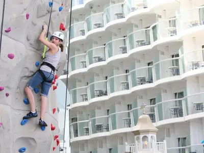 Eine Frau klettert an einer Wand, im Hintergrund die charakteristischen Balkone der Allure of the Seas, ausgestattet mit Tischen und Stühlen.