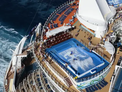 Blick auf das Oberdeck der Allure of the Seas. Ein FlowRider Surfsimulator mit Surfern ist umgeben von Passagieren und Liegestühlen. Links ist das offene, wellige Meer zu sehen.